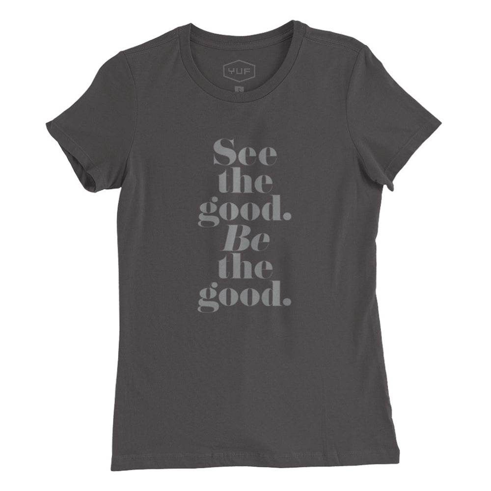 A women’s cut fashion t-shirt in Asphalt Gray, with elegant typography in a vertical stack: “See the good. BE the good.” By fashion brand YUF, for wolfsaint.net