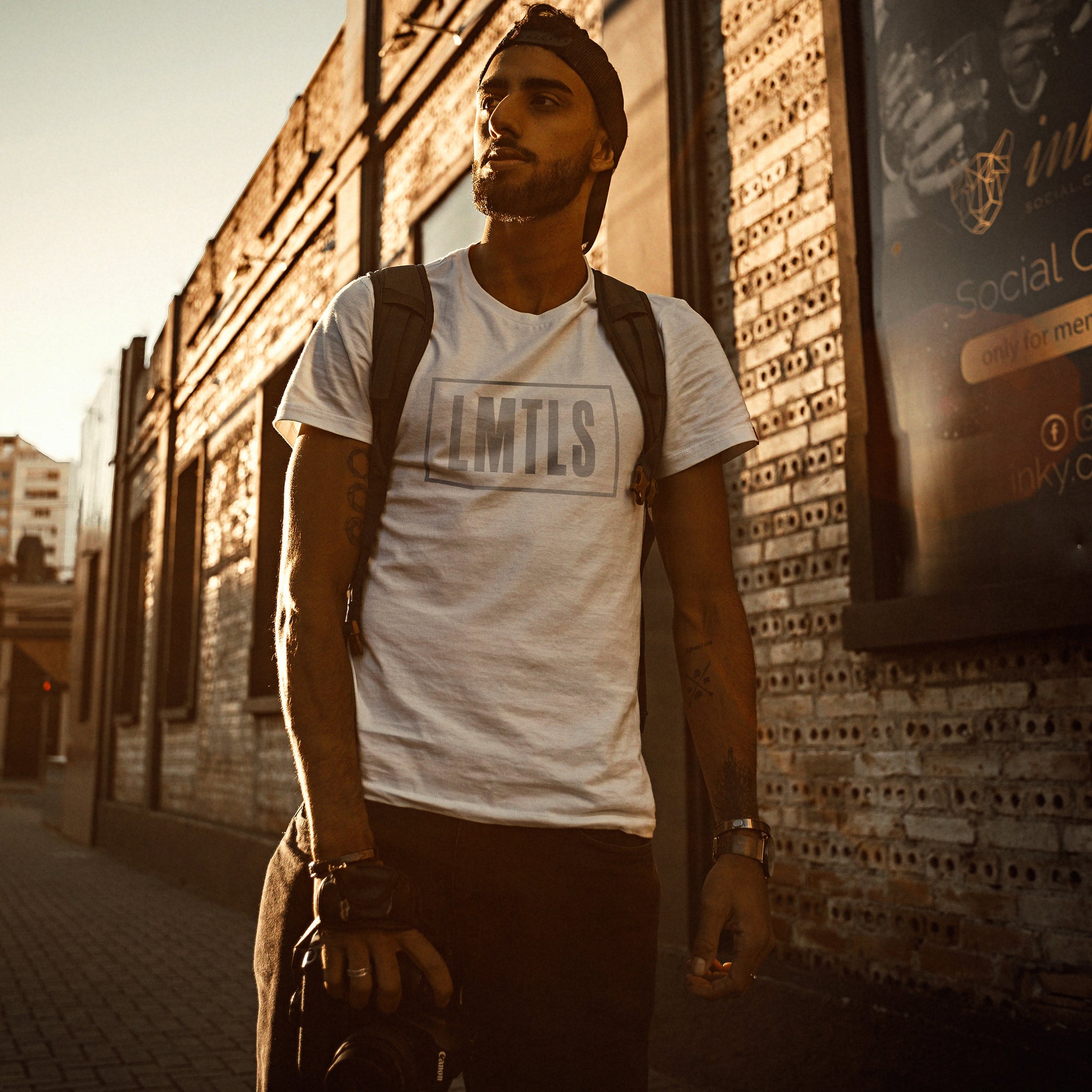 A young man with his camera, wearing an inspirational, motivational graphic t-shirt in White, with the typographic treatment LMTLS, representing a “limitless” lifestyle. On the back of the shirt is the number 10, as in a 10 out of 10, perfect score. By fashion brand YUF, from wolfsaint.net