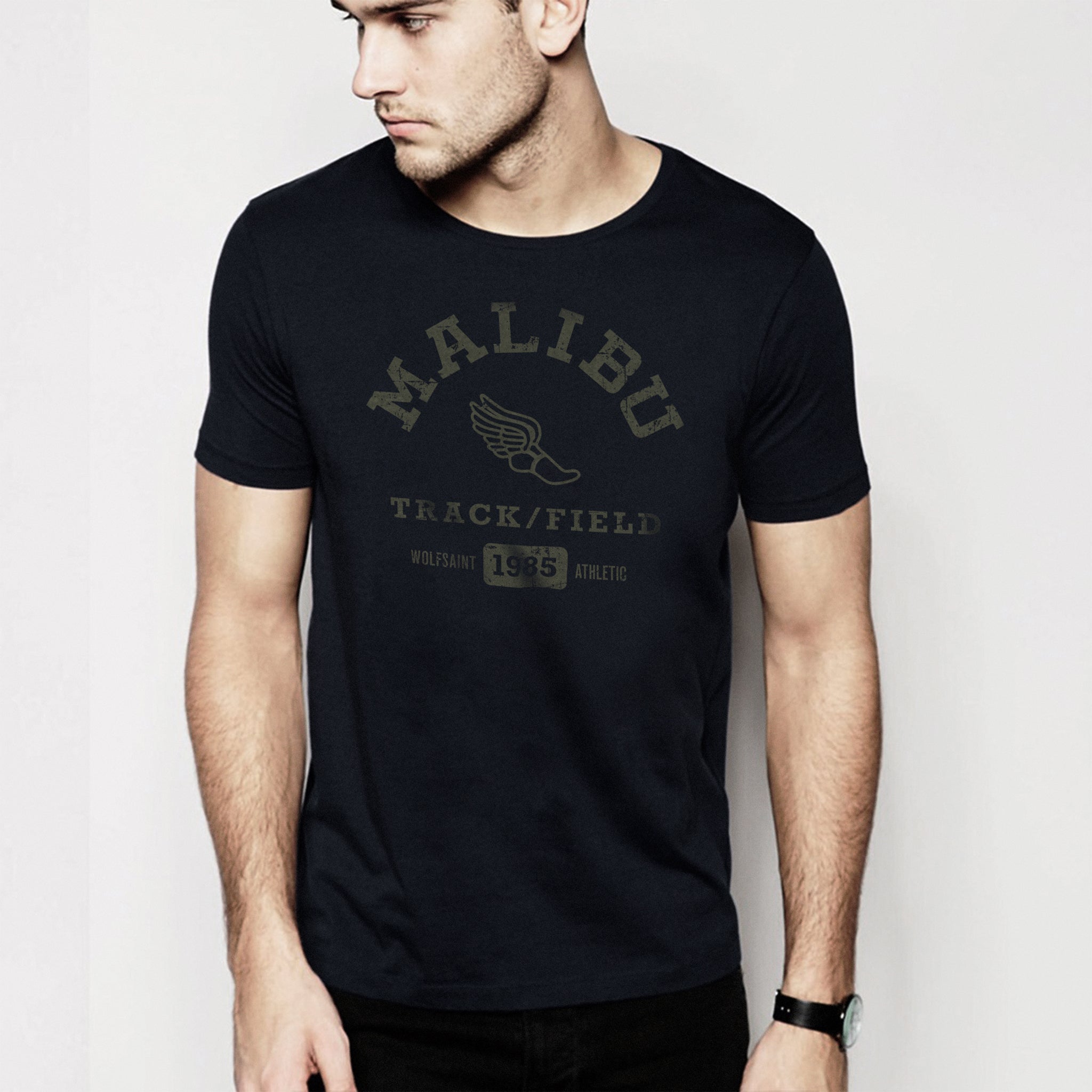 A male model in a studio wearing fashionable, vintage-inspired retro t-shirt in Classic Black, featuring a graphic representing a sarcastic and fictitious Malibu (California) Track and Field team. From wolfsaint.net