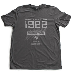 A vintage-inspired t-shirt in asphalt gray, featuring retro digital font reading “1982” with French taglines and a 45 record adapter graphic, promoting a fictional Monte Carlo discotheque from the 1970s and 80s. By fashion brand Ruston/Bond, from wolfsaint.net