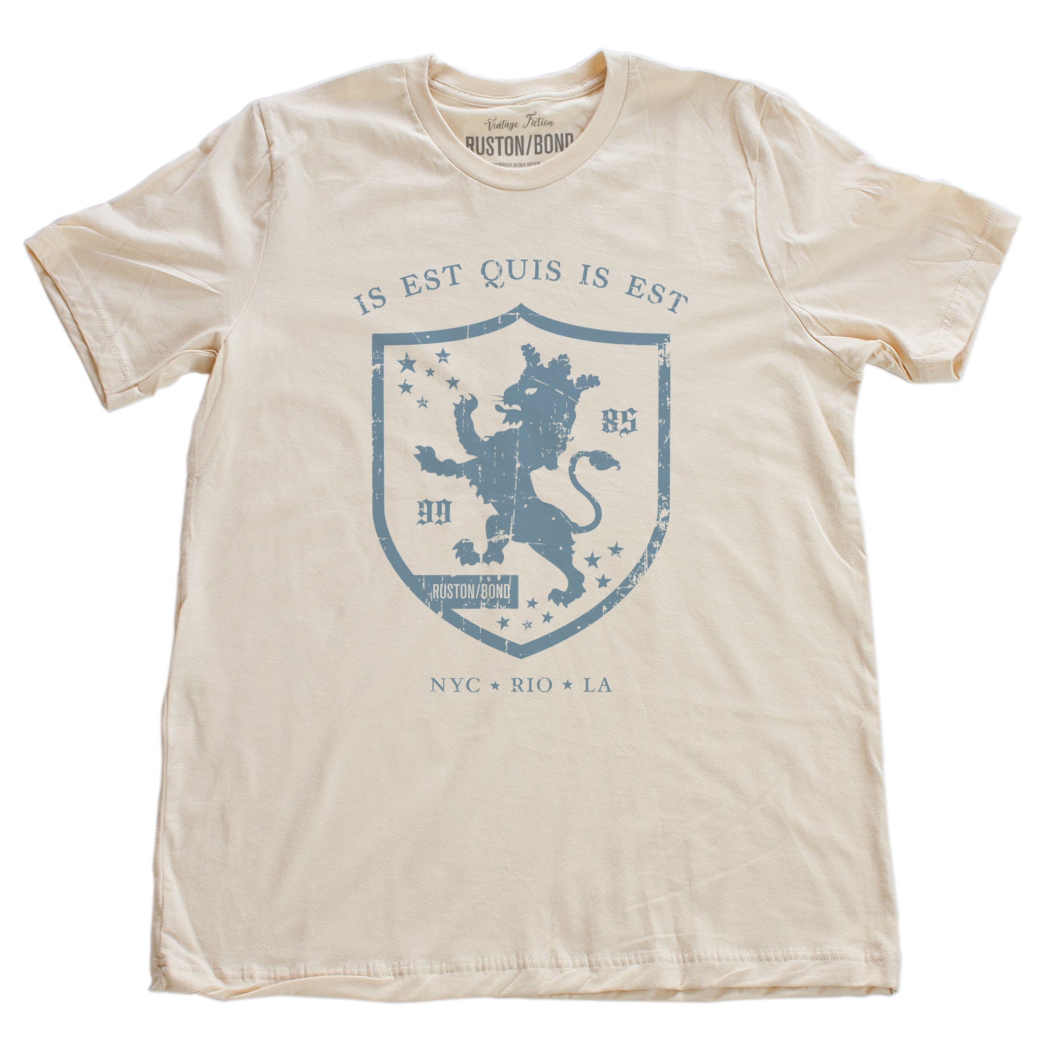 A retro, vintage-inspired fashion t-shirt in Soft Cream, with an medieval graphic of a lion within a shield,surrounded by the sarcastic text in Latin “it is what it is,” and New York, Rio, Los Angeles at the bottom. By fashion brand Ruston/Bond, from wolfsaint.net