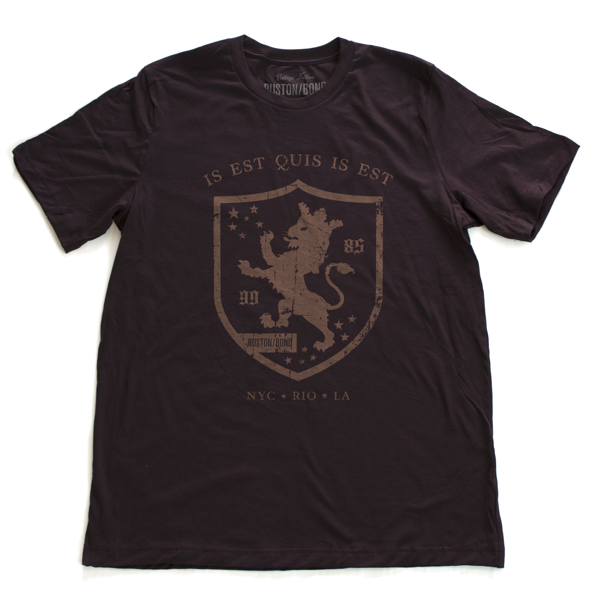 A retro, vintage-inspired fashion t-shirt in Oxblood, with an medieval graphic of a lion within a shield,surrounded by the sarcastic text in Latin “it is what it is,” and New York, Rio, Los Angeles at the bottom. By fashion brand Ruston/Bond, from wolfsaint.net