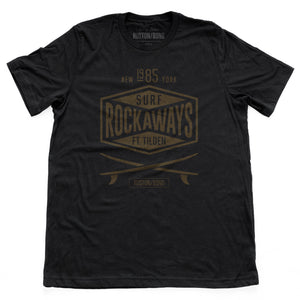 A vintage-inspired, retro design graphic t-shirt with two crossed surfboards below a typographic treatment with the words “SURF ROCKAWAYS FT. TILDEN / New York 1985” by fashion brand Ruston/Bond, for wolfsaint.net