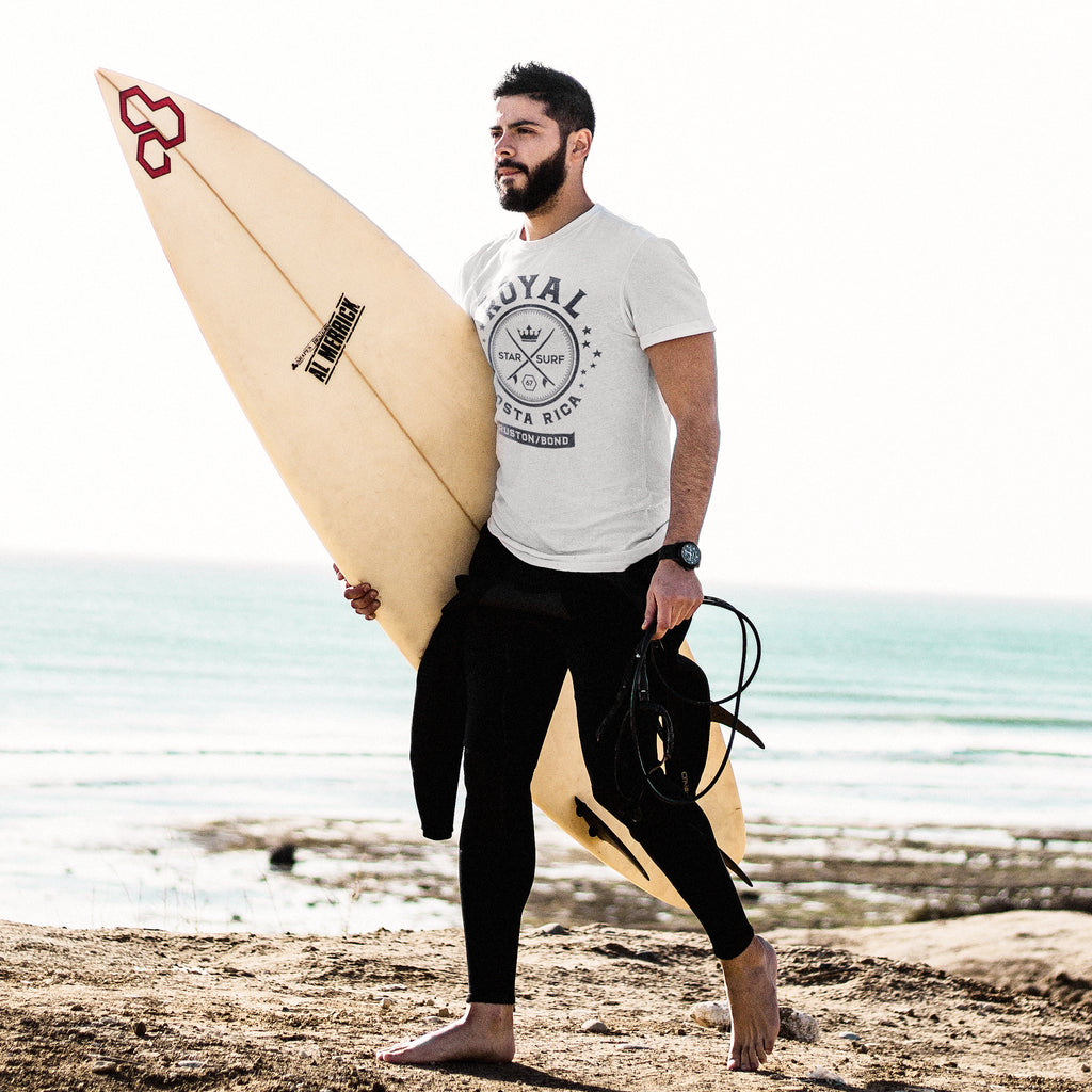 A bearded man carries a surfboard along a beach. He wears a wetsuit and a vintage-inspired White t-shirt with a retro graphic of crossed surfboards and a crown, surrounded by the words ROYAL / STAR SURF / COSTA RICA. By the fashion brand Ruston/Bond, for Wolfsaint.net