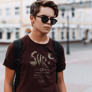 A GIF showing various colors of a fashionable graphic t-shirt with a retro, vintage design. A teenage male wearing sunglasses wears this shirt on a city street. The shirt features a surfboard graphic beneath large painted type which reads “SURF” and below it “Brazil, Rio de Janeiro, and Tamarindo, Costa Rica” above the Wolfsaint logo.  For wolfsaint.net