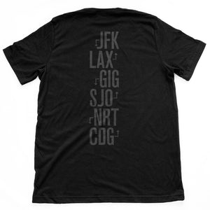 The back of the black version of a fashion t-shirt representing a jetsetting lifestyle. On the front of the shirts is a simple graphic of a commercial airliner superimposed on a globe, and the word “Worldwide.” On the reverse is a stack of airport codes representing international travel (New York/JFK, Los Angeles/LAX; Rio de Janeiro/GIG; San Jose Costa Rica/SJO; Tokyo/NRT; Paris/CDG). By fashion brand WOLFSAINT, from wolfsaint.net
