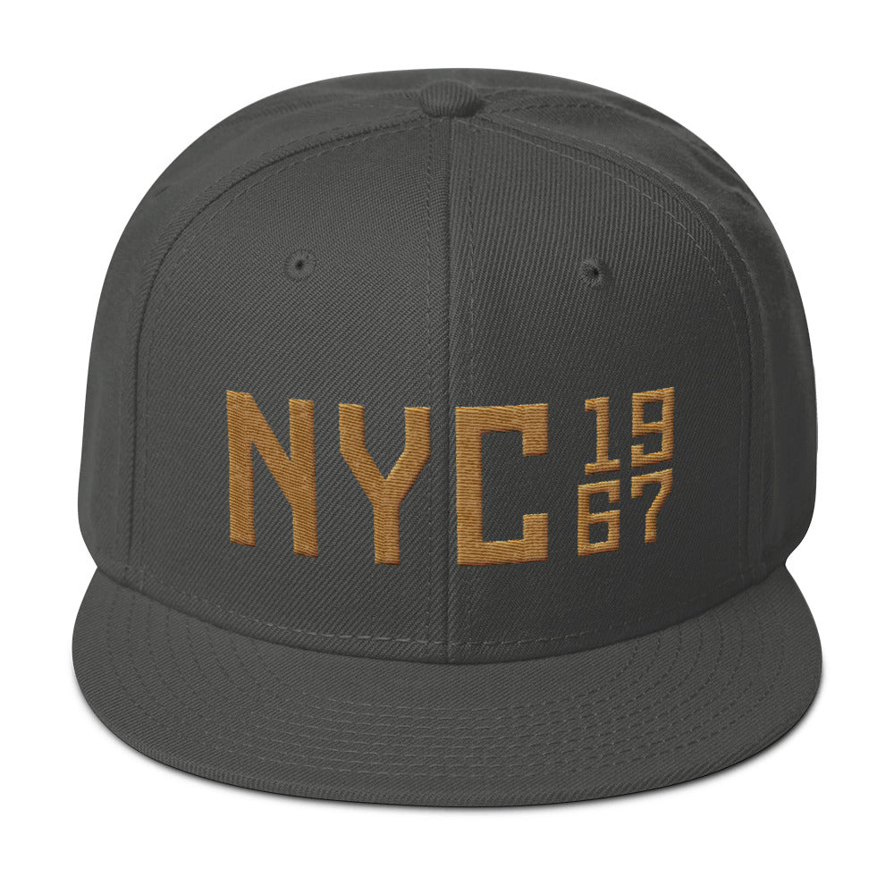 A puff embroidery high-profile SnapBack cap in Gray with Gold thread of a large “NYC” (New York City) with the vintage year 1967 in a stack to the side. From wolfsaint.net 