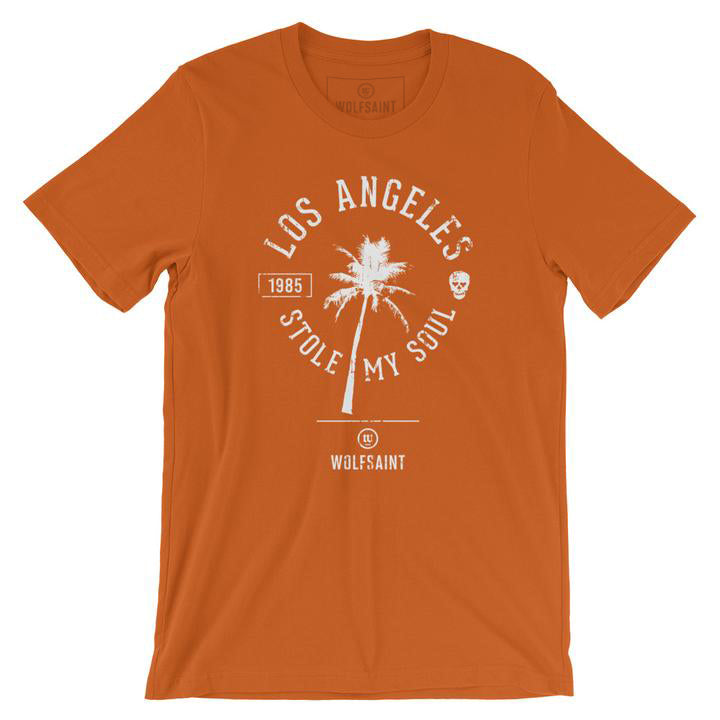 A fashionable retro graphic t-shirt in orange, featuring a palm tree surrounded by the sarcastic words “Los Angeles Stole My Soul” with the year 1985 and a fun skull icon. From wolfsaint.net