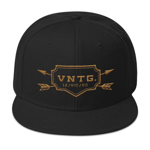 A classic SnapBack cap in Black, featuring the VNTG. logo and it’s cities (Los Angeles, New York City, Rio de Janeiro) within a shield, edged with crossing arrows. From wolfsaint.net