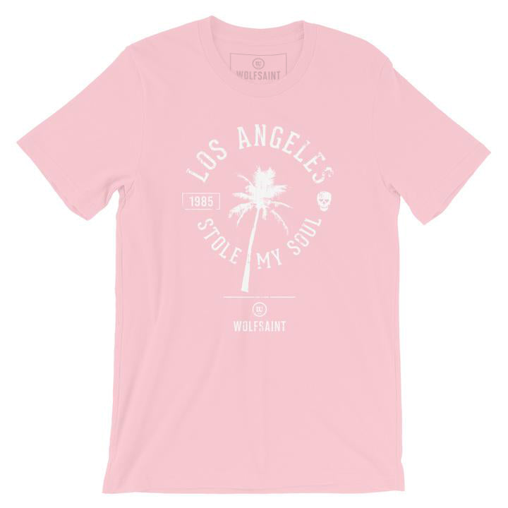 A fashionable retro graphic t-shirt in pink, featuring a palm tree surrounded by the sarcastic words “Los Angeles Stole My Soul” with the year 1985 and a fun skull icon. From wolfsaint.net