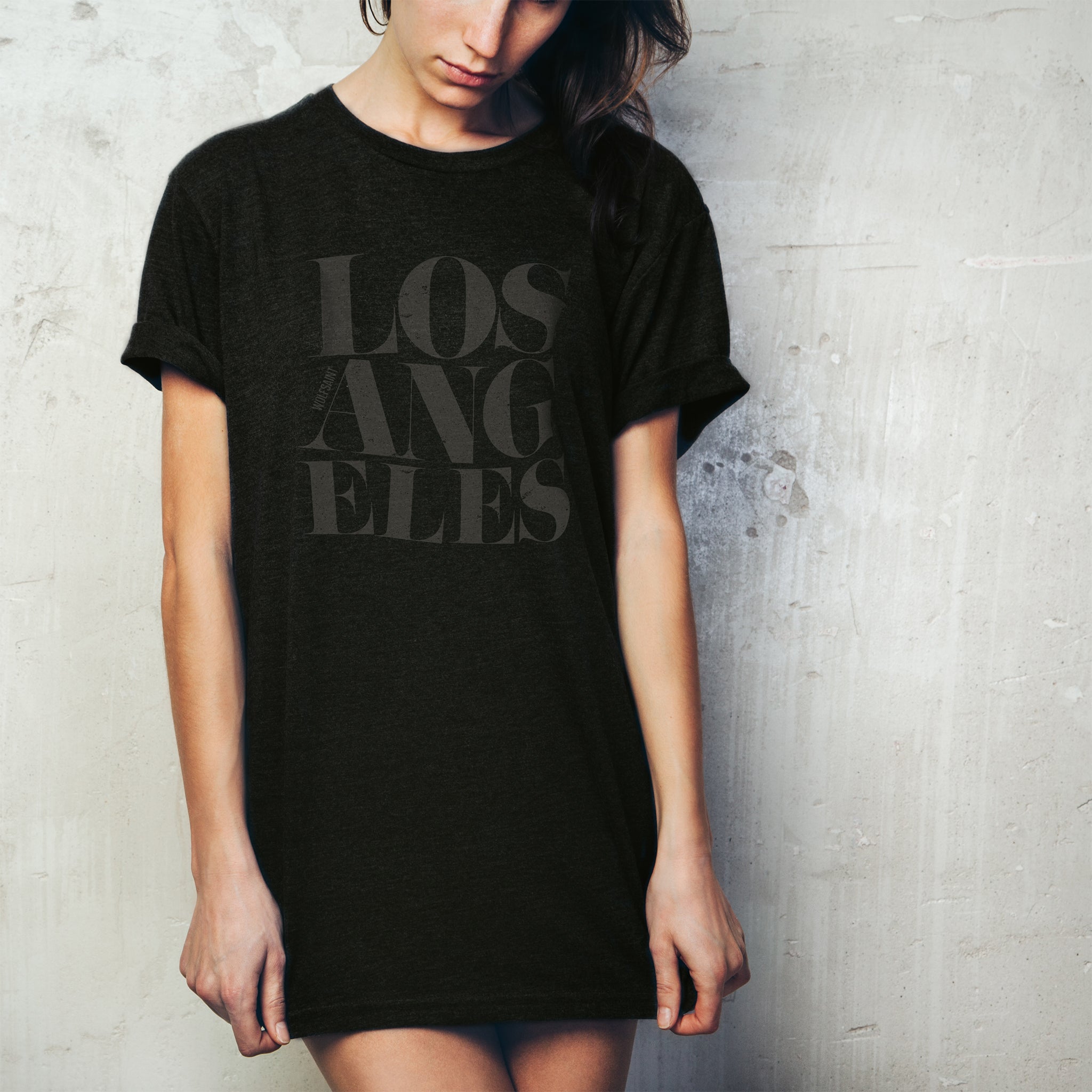 A stylish young woman wearing a fashionable black t-shirt as a shirt dress, with the word “LOS ANGELES” in a elegant font, stacked in three tilted lines to sarcastically simulate an earthquake. From wolfsaint.net