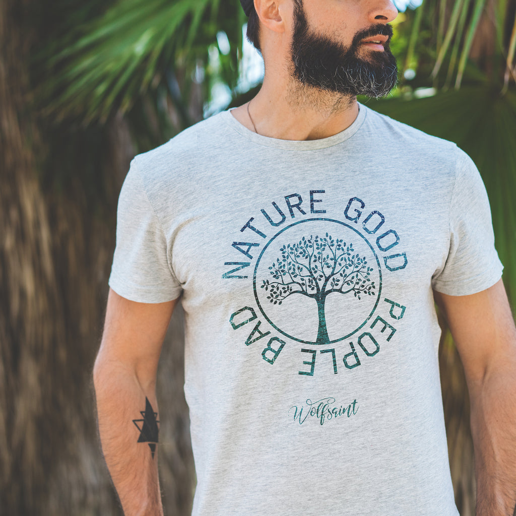 A bearded young man standing in front of palm fronds, wearing a fashionable vintage-inspired, retro t-shirt in Athletic Heather Gray, featuring a graphic of a thriving tree in a circle, surrounded by the sarcastic text: “NATURE GOOD, PEOPLE BAD” with the Wolfsaint brand script logo beneath. From wolfsaint.net