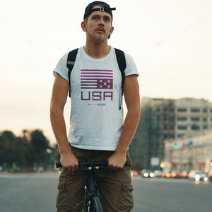 A young hipster dude on a bicycle wears a classic graphic t-shirt featuring a political statement about the American democracy, showing an upside down simplified American flag with Xs instead of stars, above the letters “USA” and the WOLFSAINT gothic logo. From wolfsaint.net