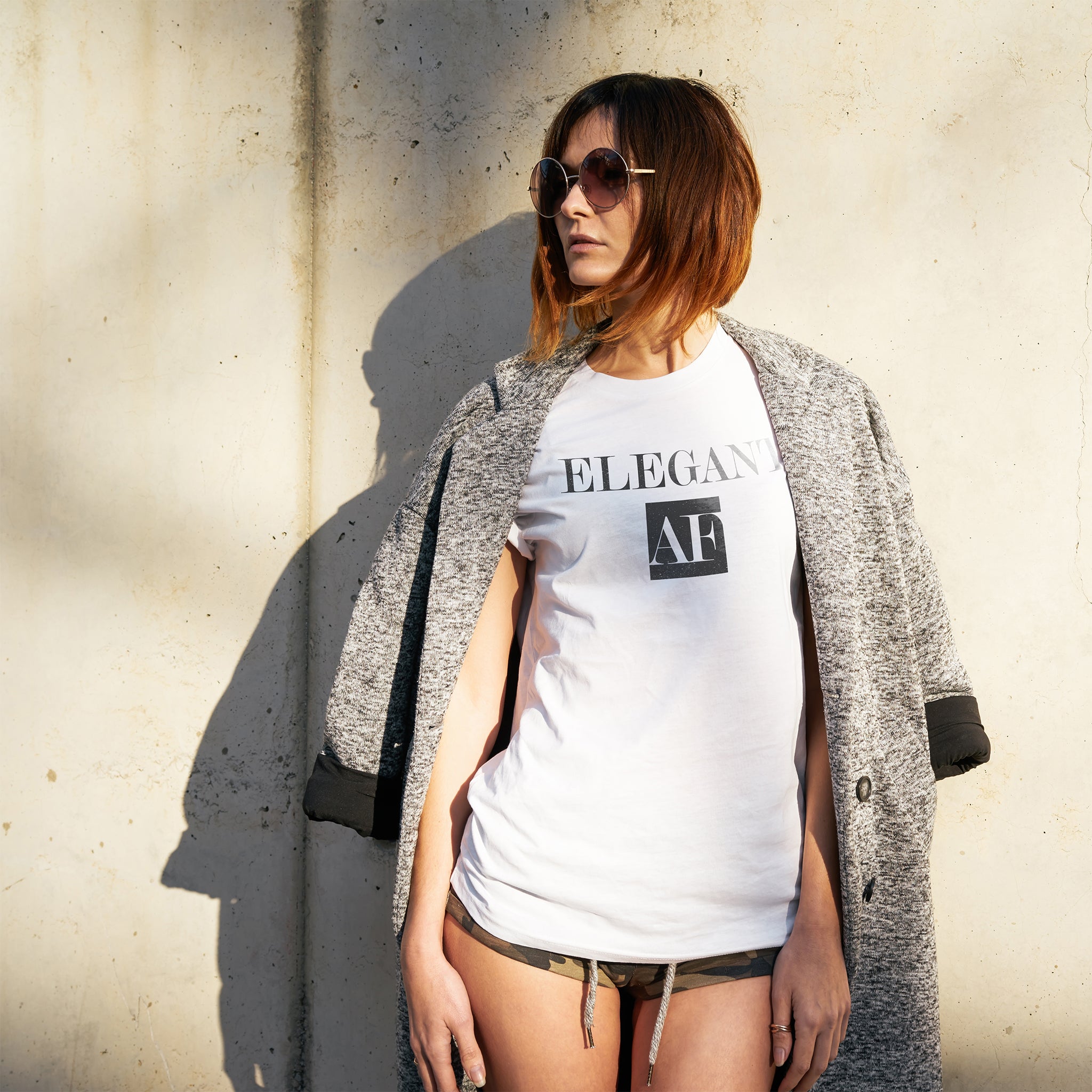 A fashionable young woman wears a stylish t-shirt with the sarcastic, meme-influenced typographic “ELEGANT AF” under a coat, in dappled sunlight. By fashion brand YUF, from wolfsaint.net
